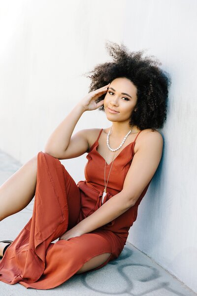 Teen girl sitting against a light wall in a rust-colored maxi dress, exuding effortless beauty with natural curls and layered jewelry in a relaxed, boho-chic pose.