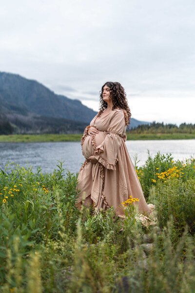 A pregnant women in a beautiful pink dress is surrounded by wildflowers and views of the Columbia River Gorge and Columbia River in Washington State.