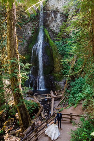 a couple in wedding attire dancing below Marymere Falls in Washington.
