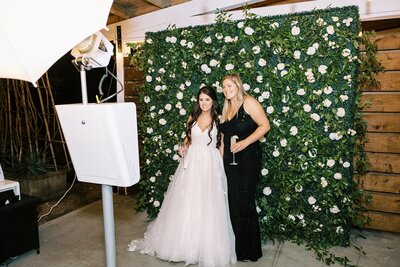 a bride poses with a woman in a black dress in front of a flower wall in an open air photo booth at her wedding.