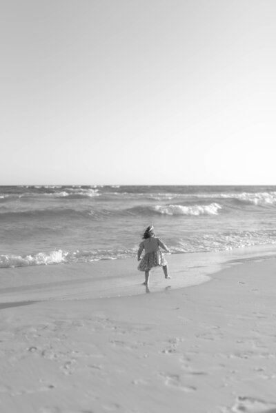 A black and white photo of a little girl at grayton beach in 30A Florida dancing and jumping as the waves come up to shore. 