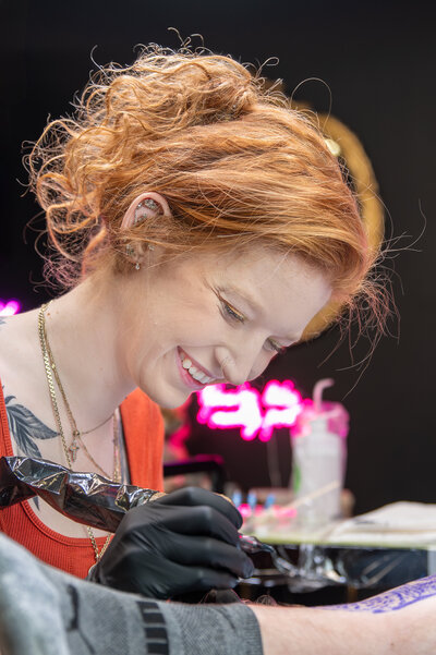 Tattoo artist wearing an orange shirt at local Ottawa studio smiling with pride for her work as she tattoos a client