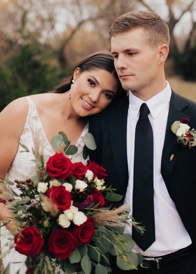 Bride lays head on grooms shoulder holding a bouquet full of roses and her timeless hair slicked back while the groom is wearing a timeless black and white suit at the  Highlands venue in wichita, kansas