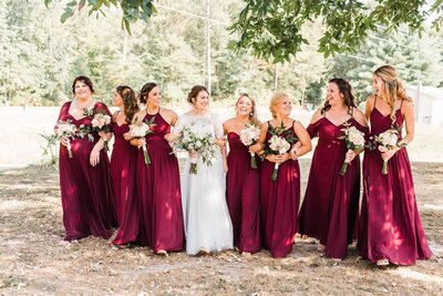 bride in white dress with sleeves and bouquet walking with bridemaids in maroon dresses in a field coordinated by cait and co events