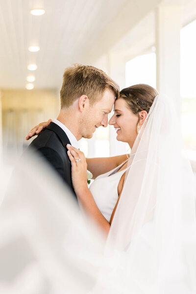 Bride and groom on a white porch with foreheads touching as the bride's veil  comes towards the camera by Kansas City Wedding Photographer Sarah Riner Photography.