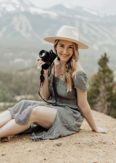 Colorado wedding photographer, Nicole DeBruyne, sits on a mountain ledge holding her camera with a mountain landscape in the background.