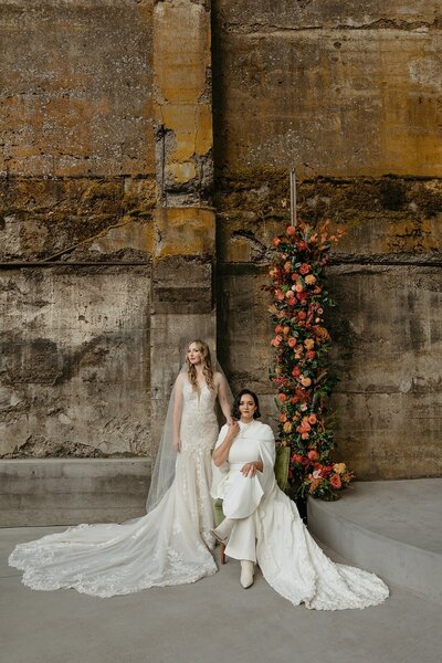 Brides Posing with Green Velvet Chair & Floral Ceremony Backdrop - Megan & Amber | Hood River Wedding  - LGBTQ Wedding