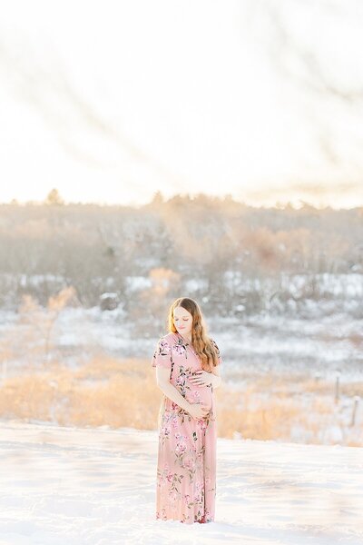 pregnant mom stands in snow during winter maternity session  with Sara Sniderman Photography  at Medfield State Hospital in Medfield Massachusetts