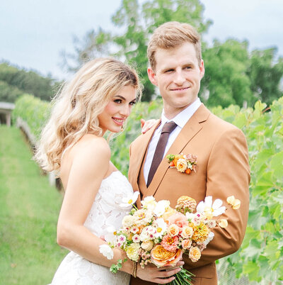 A beautiful wedding photo of a smiling bride in a strapless lace gown holding a pastel bouquet, standing beside her groom in a tan suit with a matching boutonniere, set against a lush vineyard backdrop. Perfect for capturing love and elegance on a special day.