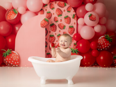 one year old girl sitting in tub laughing holding a strawberry for cake smash photoshoot