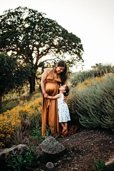 mom in gold maternity dress looks down at belly in a field of wildflowers