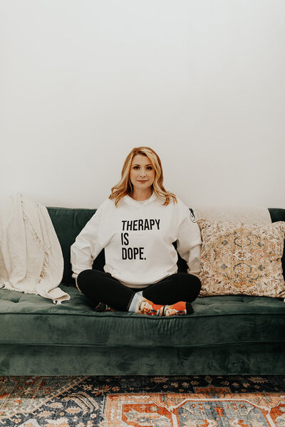 Shylah faces the camera, sitting on a green velvet couch, surrounded by throw pillows and comfy blankets. She is sitting cross-legged, wearing socks with Mr. Rogers' face, as well as a white hoodie that says "THERAPY IS DOPE."