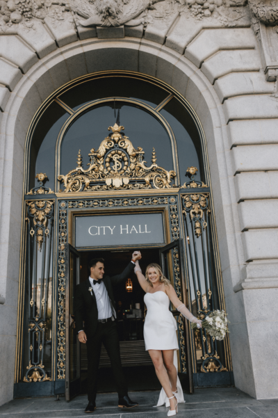 A joyful newlywed couple exits a grand building through ornate golden doors marked "City Hall" in San Francisco. The bride, in a white dress, playfully raises her bouquet as the groom holds her hand