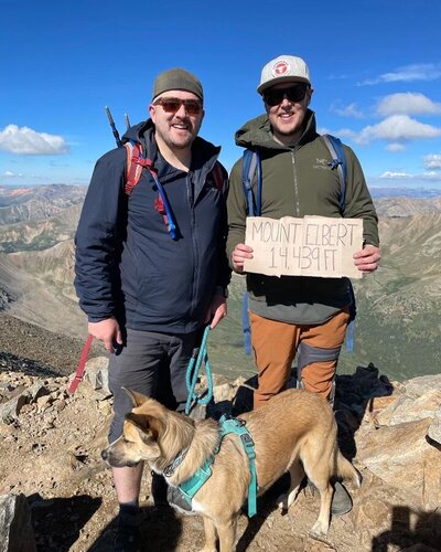 two men with dog in hiking clothes with sign 'mount elbert 14,439ft'