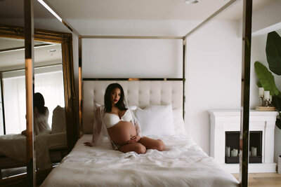 Pregnant woman in white lingerie sitting on bed for a maternity boudoir photoshoot