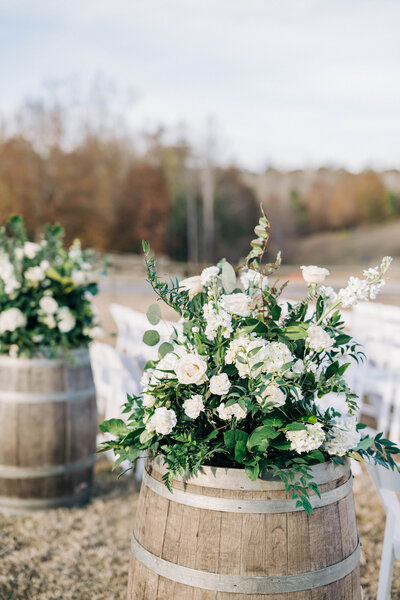 wooden barrel with green and white floral centerpiece on it and white chairs behind it in rows