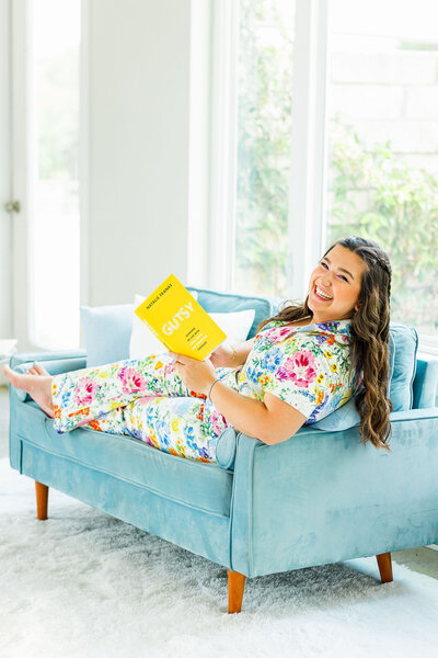 Makayla Rivera wearing a colorful jumpsuit sitting on a blue couch with a yellow book
