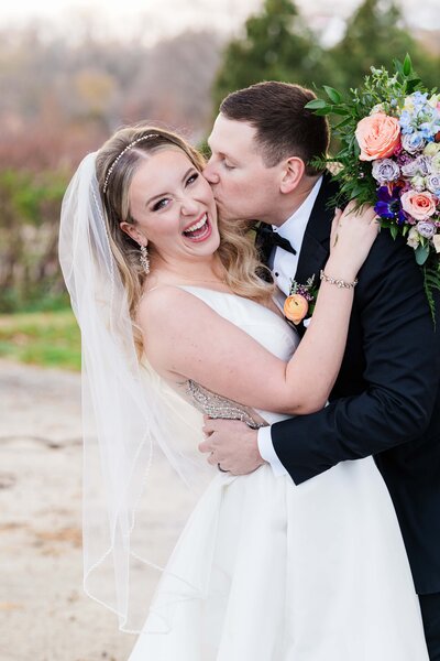 A bride and groom joyfully embracing and kissing on their wedding day, with the bride holding a colorful bouquet, thanks to their stress-free wedding package.