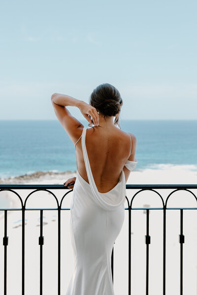 Bride in an open-back wedding gown admires the ocean view from her balcony suite at Acre Resort, Cabo.