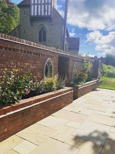 Red brick garden wall with arched stone insets and raised flower beds, set beside a sandstone patio with a historic stone church in the background.