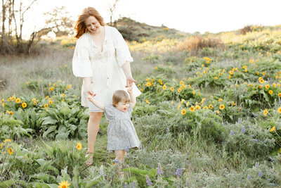 Mama in cream peasant dress smiling down at her one year old baby girl who is holding her hands while she walks. Baby is wearing a grey dress with white flowers and is smiling with her tongue sticking out. They are wlaking through a field of yellow wildflowers at sunset.