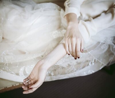A womans hands with painted red nails are laying folded on top of her white dress