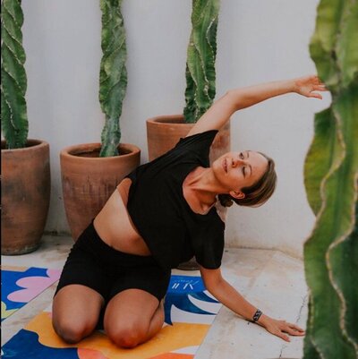 Nicole Peter, a yoga instructor in Tulum, practices a side-body stretch on a vibrant yoga mat, surrounded by potted cactus plants in a tranquil outdoor setting.