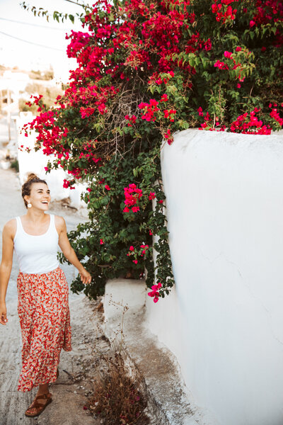 Katie Arnold, destination wedding photographer, walking by bright red flowers in a Mediterranean village.
