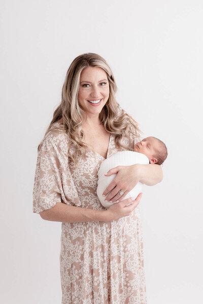 Light-skinned mom in beige floral dress holding newborn baby wrapped in white swaddle. Woman is smiling at camera. Baby is sleeping.