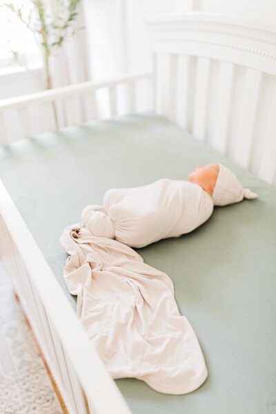 Newborn boy in cream swaddle and hat lies in white crib with sage green bedding taken by Little Rock newborn photographer Bailey Feeler Photography