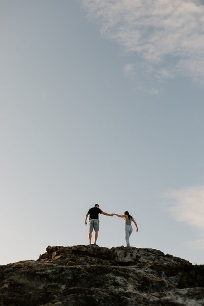 A couple holding hands as they stand at the top of a large rock 