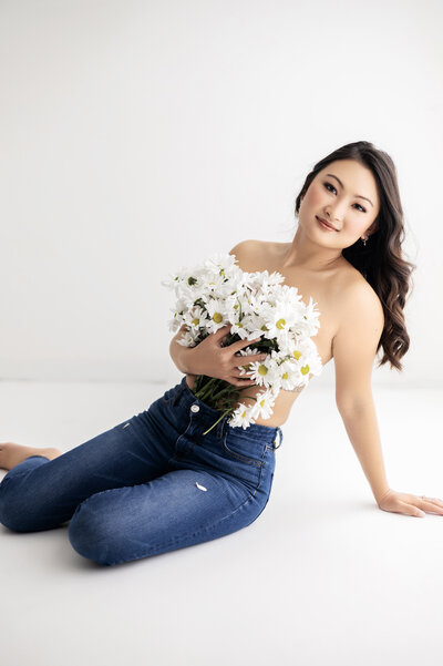 bridal boudoir portrait woman sitting in white studio holding white daisy's over chest in blue jeans