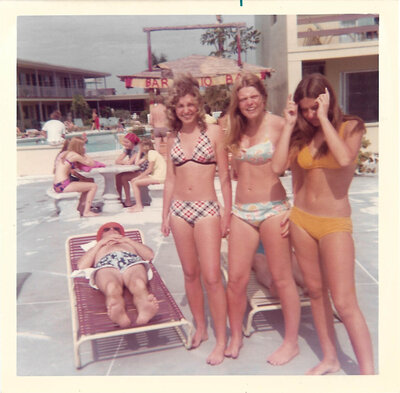 three women in bathing suits stand behind a man who is sleeping and sun tanning, in the 1970s.