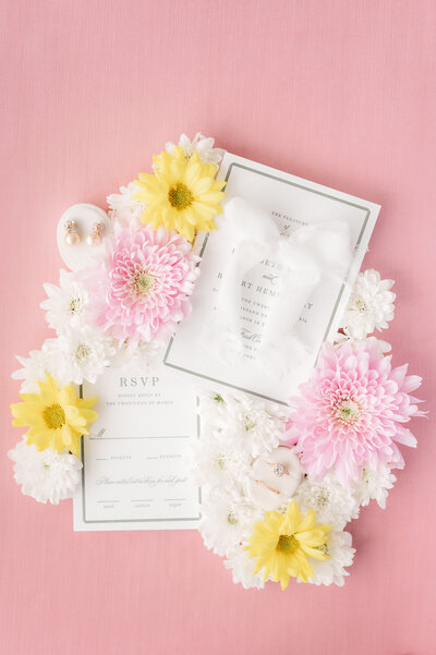 wedding invitations on pink backdrop with white, pink, and yellow flowers around them