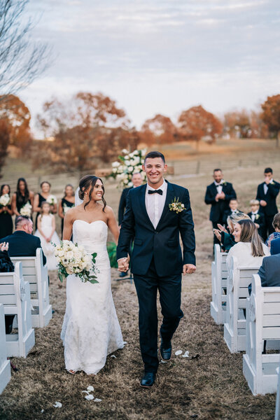 woman in white dress and man in black tux walking down an aisle with rows of white chairs