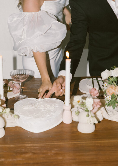 a bride and groom photographed on 35mm film with pastel details as the tablescape and dipping their fingers in a heart shaped cake that says "lovers" in white icing