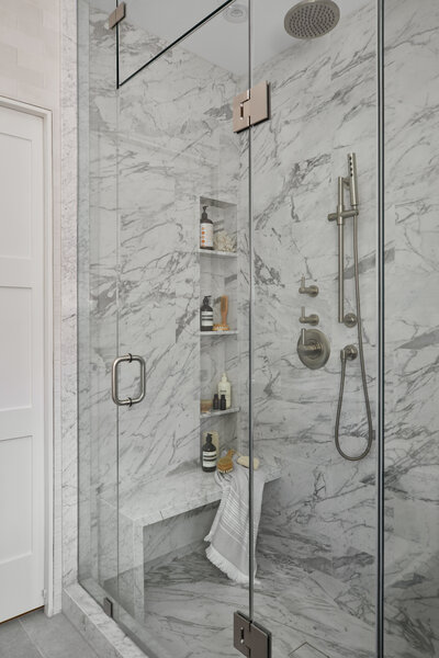 A luxury marble walk-in shower with built-in shelving and a bench, brushed nickel accents, and a sliding glass door
