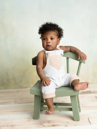Smiling baby in a white dress and headband sits on an ornate, padded chair. The setting is soft and warm, conveying a joyful and gentle atmosphere.