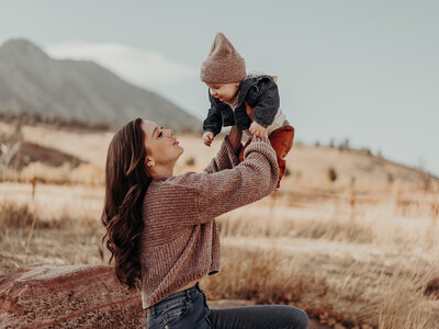 A gorgeous brunette mother holds her six month old, baby boy in the air, smiling at him with a beautiful scenic mountain in the background during fall.