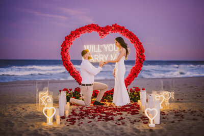 man on his knee during proposal on the beach at sunset in front of a floral heart arch at long branch nj