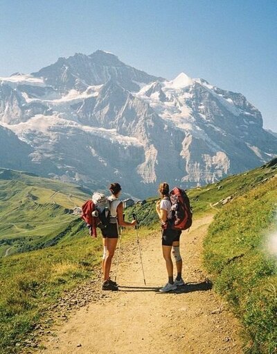 two women with backpacking gear stopped on a hiking trail and looking at a big mountain
