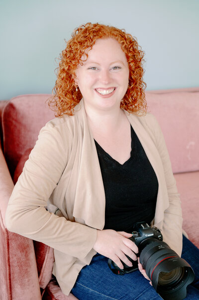 Val smiling in a black shirt and tan cardigan while holding a camera