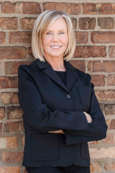Woman with blonde hair wearing a black blazer stands confidently with arms crossed in front of a brick wall, smiling at the camera.