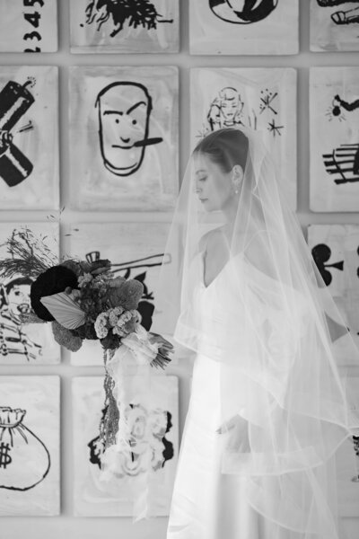 Black and white photo of a bride holding a unique, textured bouquet, wearing an elegant veil and gown, standing against a backdrop of abstract modern art.