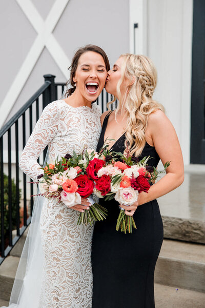 Bridesmaid wearing black dress kissing bride on cheek while they hold bright red and white garden style bouquets