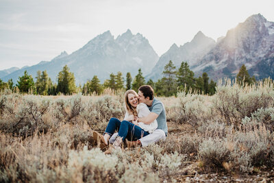 Jackson Hole wedding photographer captures engaged couple sitting in a sage brush field in the Tetons sittign together and laughing and kissing
