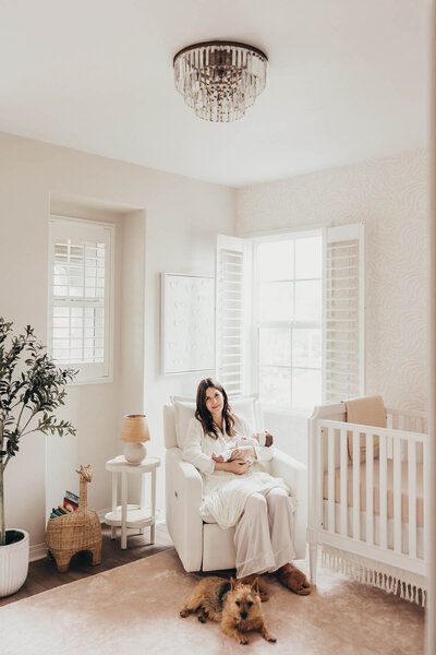 A mom dressed in white silk pajamas holds her newborn baby while she sits in her beautiful nursery in her Carlsbad home with her dog at her feet.