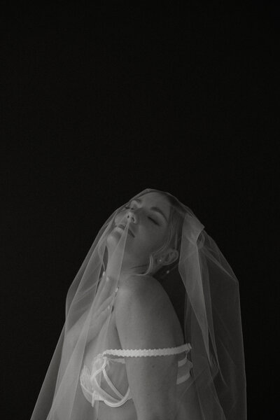 Black and white photo of woman in white lingerie and a veil tilting her head back in front of a black background