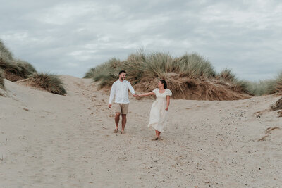 Couple hold hands walking through the sand dunes smiling during a photoshoot at Camber Sands beach, Rye