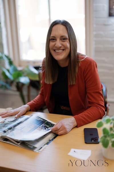 Alex is sitting at a table, flipping through a wedding magazine. Her phone and Wedding Planning business cards rest on the table beside the magazine as she smiles at the camera. She's been working.